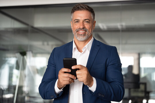 Smiling mature Latin or Indian businessman holding smartphone in office. Middle aged manager using cell phone mobile app, looking at camera. Digital technology application and solutions for business.