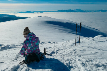 Woman sitting on snowy ground with scenic view of fog covered Lavanttal valley. Snow capped mountains Karawanks and Julian Alps seen from top of Grosser Speikkogel, Kor Alps, Carinthia Styria, Austria
