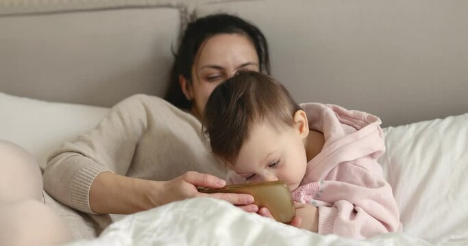 Mother and child playing with smartphone while sitting on bed in bedroom at home