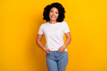 Photo of adorable cheerful woman with perming coiffure dressed white t-shirt keep arms in pockets isolated on yellow color background