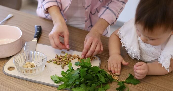 female hands cracking pistachio nut shell for preparing salad in the kitchen, child helping mother to cook