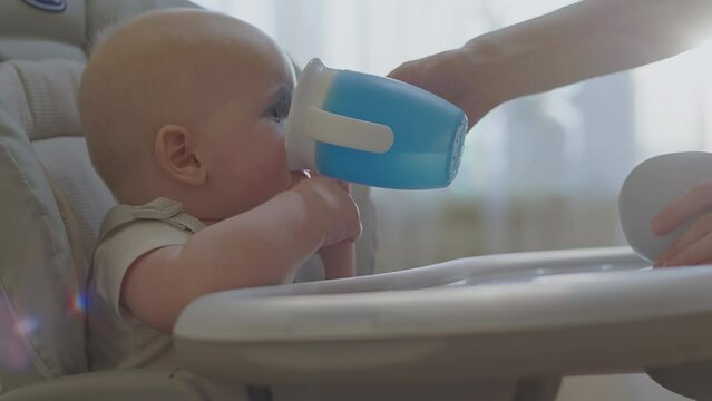 Messy face infant boy refuse sippy cup from mother hand while seat in high chair. Baby expression captured mid-refusal, emphasizing common feed challenges faced by parent. Child drink water first time