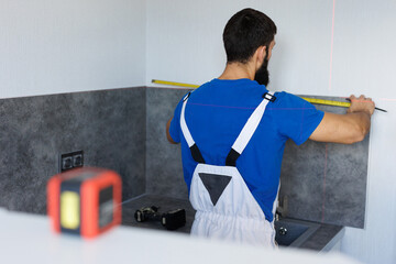 Assembler uses a tape measure and a laser level to install wall panels and cabinets in kitchens.