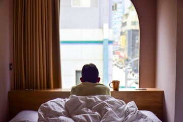 A South Asian man looking out of hotel room window admiring cityview. Selective focus, solo travelling, relaxing,  enjoying morning tea.