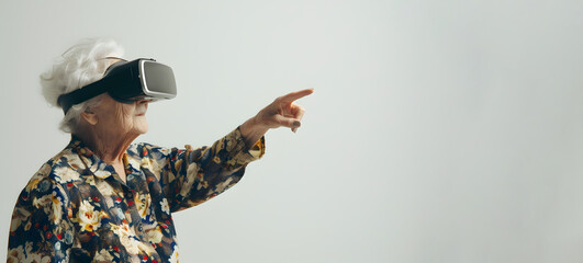 An elderly woman in VR glasses on a light plain background points her finger at an empty space for text. - obrazy, fototapety, plakaty