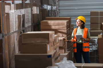 happy female warehouse worker in safety vest and hard hat holding digital tablet near cargo