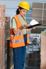 female warehouse worker in hard hat and safety vest holding clipboard checking inventory