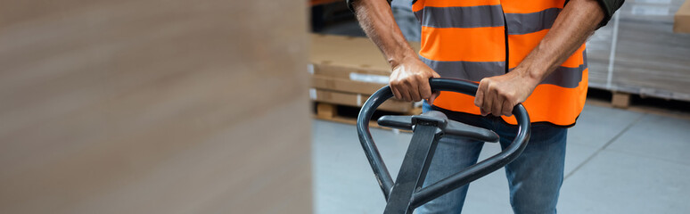 cropped banner of warehouse worker in hard hat and safety vest transporting pallet with hand truck