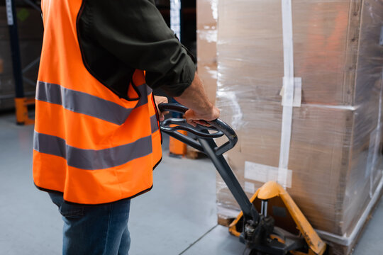 cropped middle aged warehouse worker in hard hat and safety vest transporting pallet with hand truck - Powered by Adobe