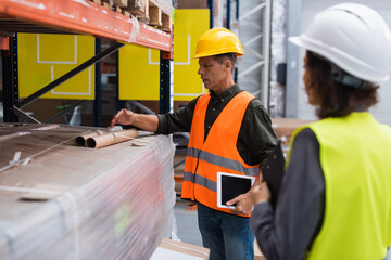 middle aged supervisor in hard hat holding tablet while explaining work to employee in warehouse