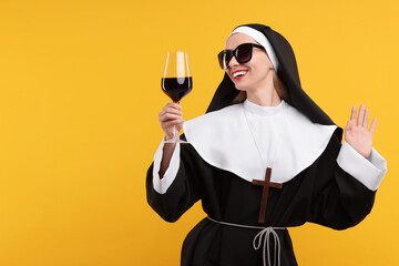 Happy woman in nun habit and sunglasses holding glass of wine against orange background. Space for...