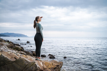 Side view of beautiful smiling young woman standing on the stone beach, traveler looks at sea in...