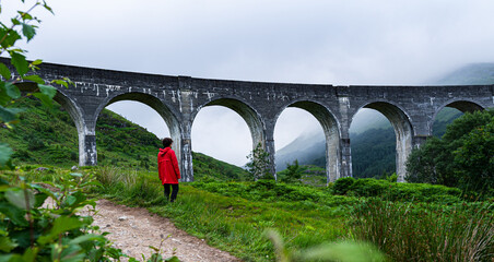 Highland Serenity: A Woman's Journey Through Scotland's Mystical Landscape, Standing Before the Stone Bridge on a Cloudy Day in her Red Coat