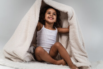 Obraz premium A little cute dark-skinned girl 2 years old plays with a fluffy white blanket on a light plain background. Cozy photos of a cheerful child wrapped in a blanket.