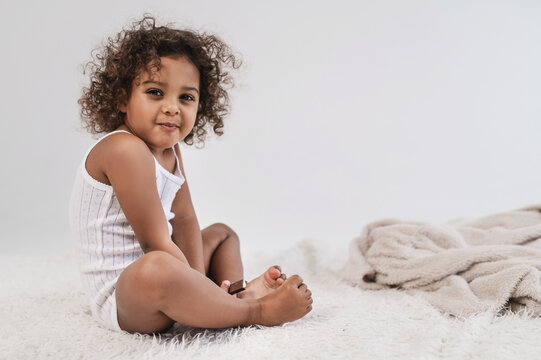 A little cute dark-skinned girl 2 years old plays with a fluffy white blanket on a light plain background. Cozy photos of a cheerful child wrapped in a blanket.