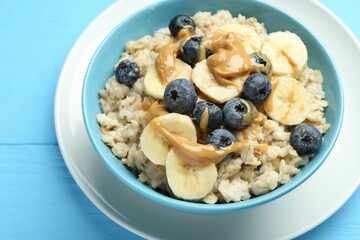 Tasty oatmeal with banana, blueberries and peanut butter served in bowl on light blue wooden table, closeup