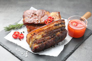 Pieces of baked pork belly served with sauce, rosemary and chili pepper on grey table