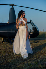 Alluring woman in a white dress, sipping champagne by a helicopter, exuding the charm and allure of...