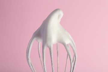 Whisk with whipped egg whites on pink background, closeup