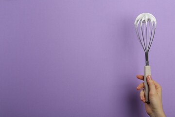 Woman holding whisk with whipped cream on violet background, closeup. Space for text