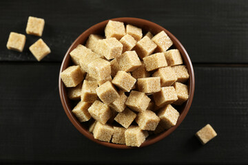 Brown sugar cubes in bowl on black wooden table, top view