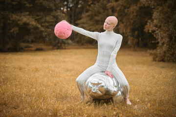 Young hairless girl with alopecia in white cloth sits on tardigrade figure and holds pink ball in hand on fall lawn park, surreal scene with bald teenage girl engages with symbolic elements - obrazy, fototapety, plakaty