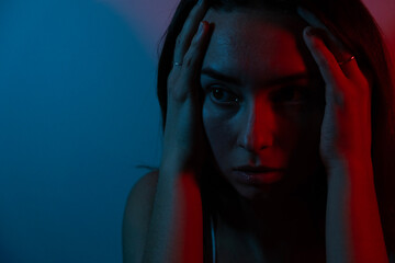 Emotional photo. Blue and red colors. Portrait photo of a woman. A young woman feels stressed and anxious.
Sad woman complaining alone sitting in the dark.Depressed young woman coping with loss, grief