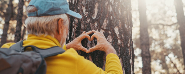 Head shot portrait close up of one old cute man taking care and protecting big tree in the forest of mountain in the nature. One mature person making a heart shape with hands loving nature concept.