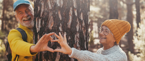 Head shot portrait close up of one old cute man taking care and protecting big tree in the forest of mountain in the nature. One mature person making a heart shape with hands loving nature concept.