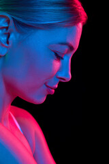 Close up photo of young attractive woman looking down against black studio background in colorful neon light. Concept of natural beauty, anti aging, cosmetology, female health. Ad