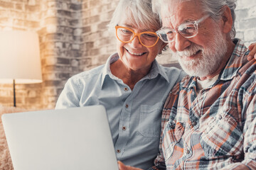 Cute couple of old people sitting on the sofa using laptop together shopping and surfing the net. Two mature people wearing eyeglasses in the living room enjoying technology. Portrait of seniors laugh - obrazy, fototapety, plakaty