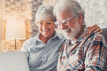 Cute couple of old people sitting on the sofa using laptop together shopping and surfing the net. Two mature people wearing eyeglasses in the living room enjoying technology. Portrait of seniors laugh