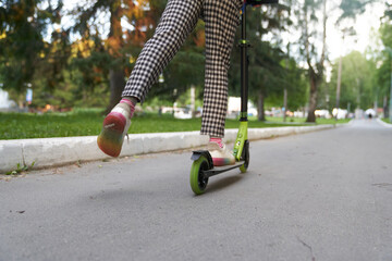 Cropped view from behind of unrecognizable girl in pants riding kick scooters on pavement on summer day, with copy space. Concept of sport, healthy and active lifestyle, eco transport, fun.