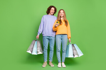 Full size portrait of two positive teenagers use smart phone hold shop bags isolated on green color...