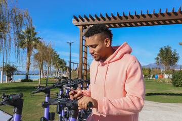 Latin guy in a pink hoodie scanning a QR code to rent an e-scooter in the park. Mixed race young man about to unlock and ride an electronic scooter. Close up, copy space, background.