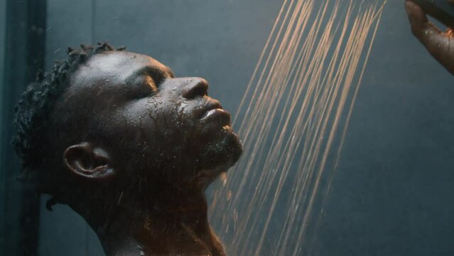 Close up African American handsome millennial 30s man washing cleaning body in dark bathroom ethnic sexy male guy taking shower bath rinsing face warm hot water morning care hygiene routine concept