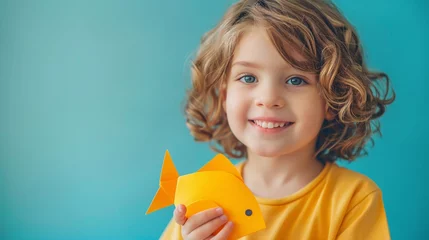 Fotobehang Enfant souriant  avec un poisson d'avril en papier sur fond turquoise » IA générative © Maelgoa