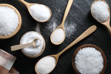 Organic white salt in bowls and spoons on black table, flat lay