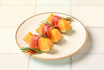 Plate of tasty melon with prosciutto and rosemary on light tile background