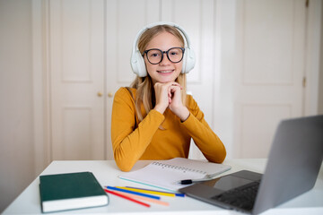 Happy teenage girl wearing headphones sitting at desk with laptop at home