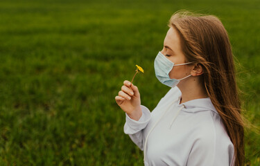 A girl in a respirator sniffs a yellow dandelion. Young blond European woman in a protective mask outdoors.