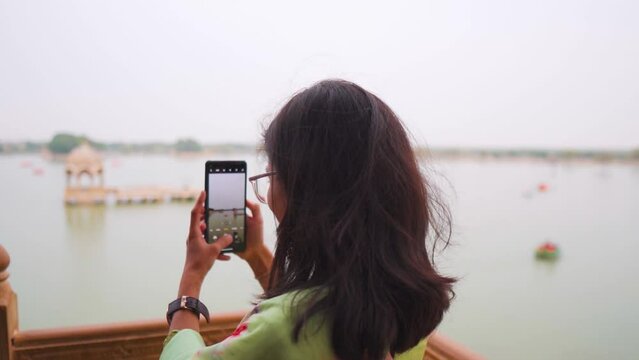 Indian tourist girl capturing video, photo of Gadisar Lake with mobile phone at Jaisalmer, Rajasthan, India. Chhatris in Gadisar Lake. Young woman capturing lake on smartphone, back view.