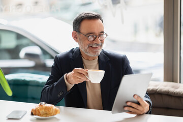 Senior man in a smart casual outfit enjoying a cup of coffee while reading on a tablet in a cafe