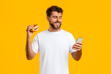 Cheerful young man holding burger using cellphone over yellow background