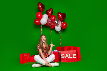 Full length photo of charming teen look directing at sale empty space sit near helium balloons presents isolated on green color background