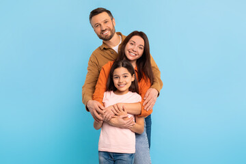 Happy parents and child daughter standing close hugging, blue backdrop