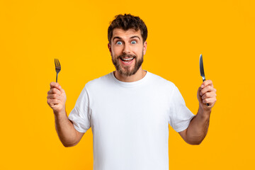 Hungry millennial man holds a knife and fork, yellow backdrop