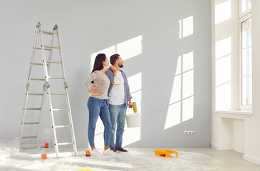 Happy married couple painting wall in their new house with paint rollers. Cheerful husband and wife...