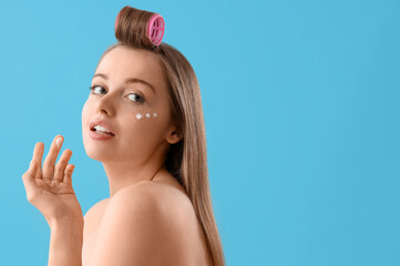 Young woman with facial cream and hair curler on blue background, closeup