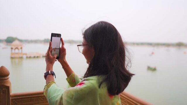 Indian tourist girl capturing video, photo of Gadisar Lake with mobile phone at Jaisalmer, Rajasthan, India. Chhatris in Gadisar Lake. Young woman capturing lake on smartphone, back view.